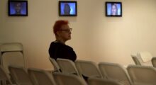 A woman with orange hair and glasses sits on a white folding chair in a gallery space.