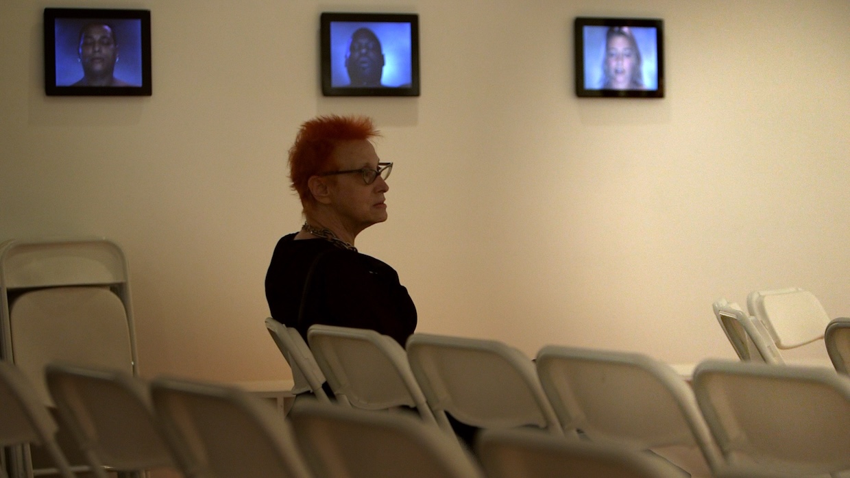 A woman with orange hair and glasses sits on a white folding chair in a gallery space.