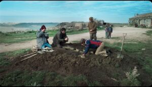 A group of men huddle around a dirt digging site on a sunny afternoon by a beach.