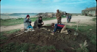 A group of men huddle around a dirt digging site on a sunny afternoon by a beach.