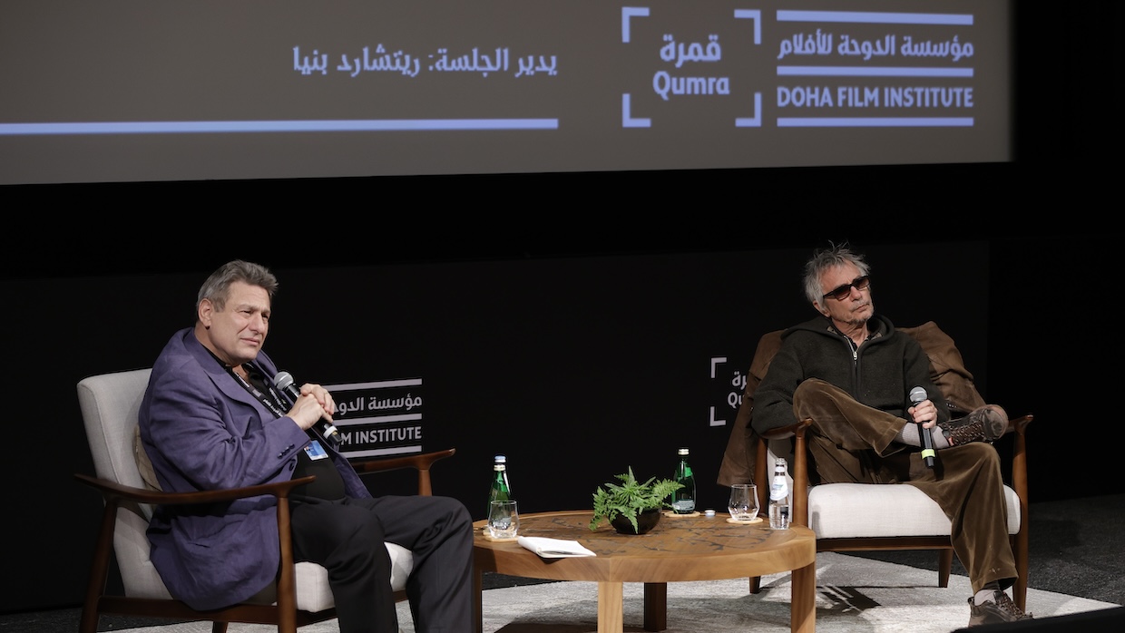Two men sit on a stage separated by a small wooden table at a Q&A.