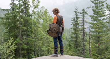 A young man holding a shield with a chipped section stands on a cliff overlooking a forest.