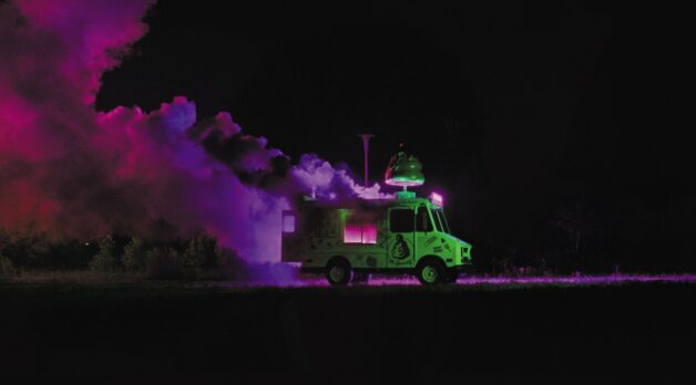 An ice cream van smokes, seemingly on fire, in a purple-lit field