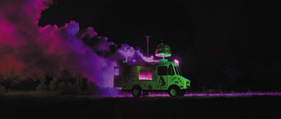 An ice cream van smokes, seemingly on fire, in a purple-lit field