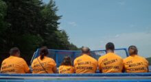 A row of people with their backs to the camera sit on a boat wearing orange t-shirts.