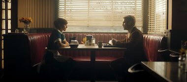 A man and a woman sit across a table looking at each other from opposite sides of a diner booth.