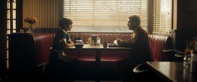 A man and a woman sit across a table looking at each other from opposite sides of a diner booth.