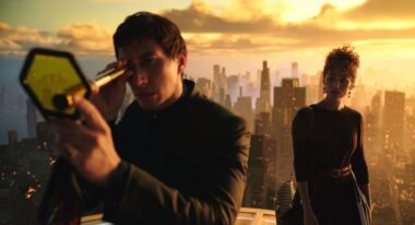 A man looks over a city landscape through a telescope at sunset while a woman stands behind him.
