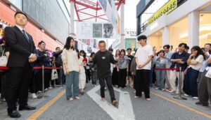 A man with bare feet walks very slowly, surrounded by an observing crowd, in Korea.