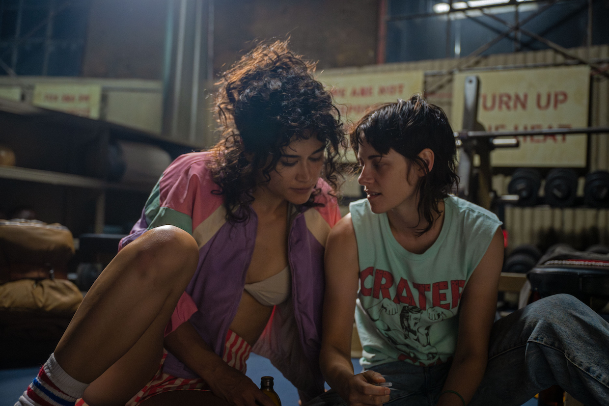 Two women lean close to each other while drinking beers and sitting on the floor of a boxing gym.