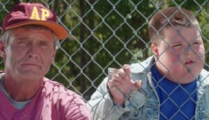 A white man and young white boy stare from behind a chain-link fence at a baseball game.