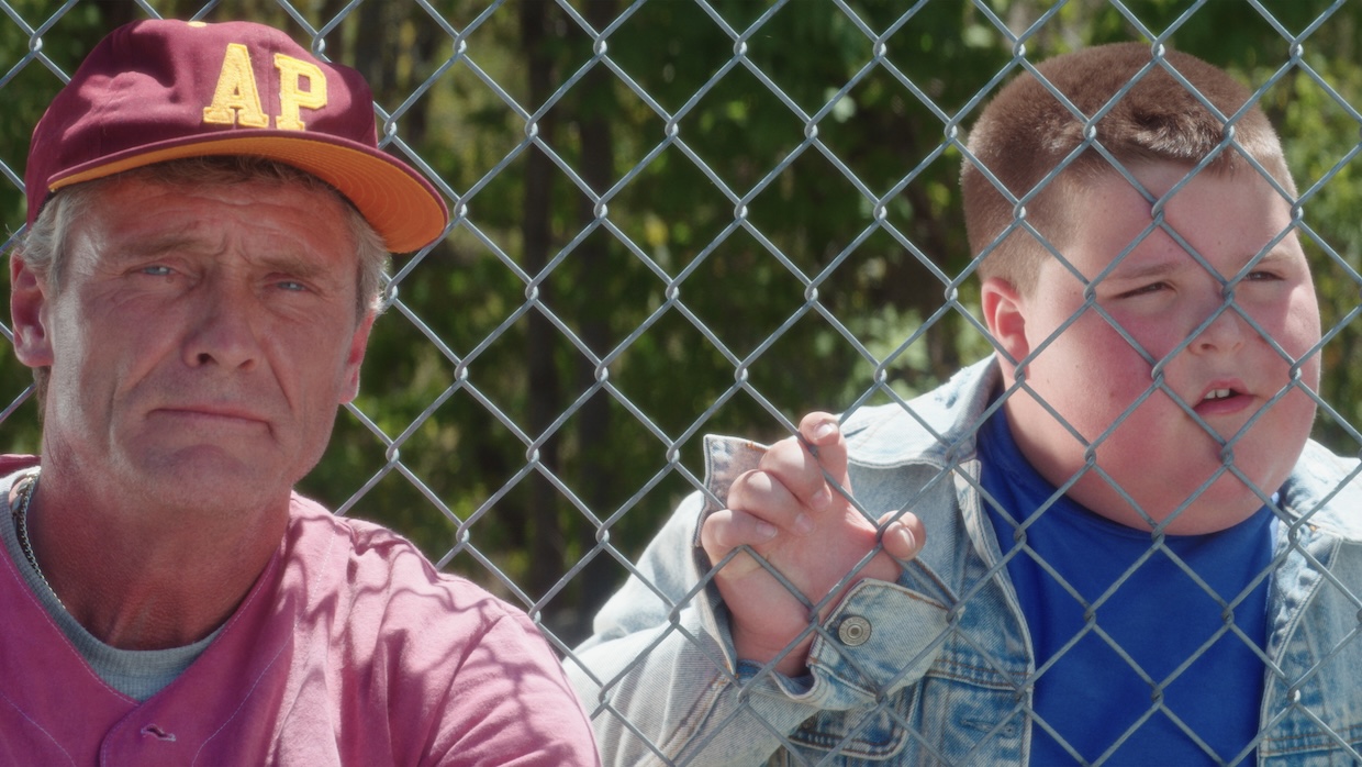 A white man and young white boy stare from behind a chain-link fence at a baseball game.