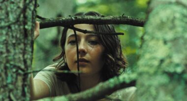 A young white teenager gazes out through tree branches in a forest.