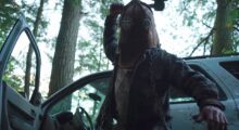 A man wearing a leather helmet strikes a blow in front of a car parked in the forest.