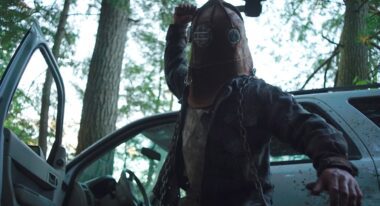 A man wearing a leather helmet strikes a blow in front of a car parked in the forest.
