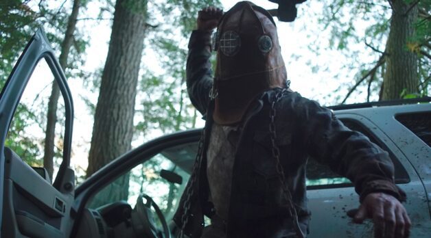 A man wearing a leather helmet strikes a blow in front of a car parked in the forest.