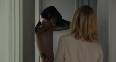 A white teenage boy pulls his t-shirt over his head in front of a woman.