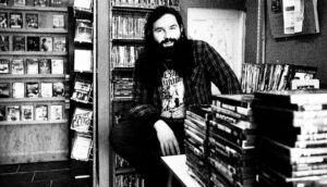 A white man with a large beard and long hair sits in a video store.