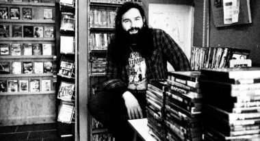 A white man with a large beard and long hair sits in a video store.