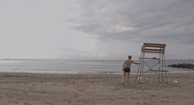 A man with his back to the camera stands facing the shoreline on a gloomy beach.