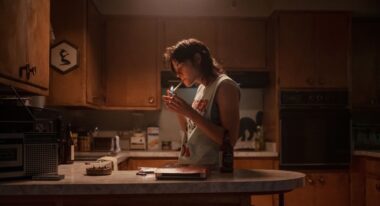A woman in a cut-off t-shirt lights a cigarette in an atmospherically lit kitchen.