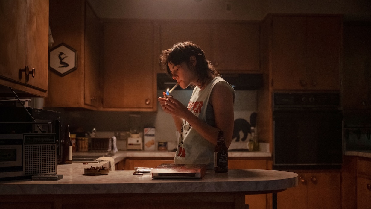 A woman in a cut-off t-shirt lights a cigarette in an atmospherically lit kitchen.