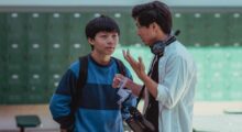 A young Asian-American teenagers listens to a 20something Asian-American director wearing headphones on a high school film set.