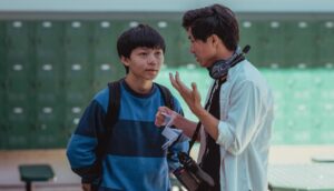 A young Asian-American teenagers listens to a 20something Asian-American director wearing headphones on a high school film set.