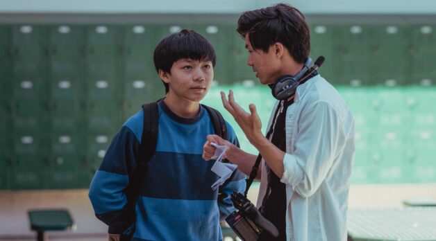 A young Asian-American teenagers listens to a 20something Asian-American director wearing headphones on a high school film set.