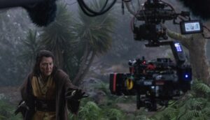 An Asian man in Jedi costume poses in front of a camera on a forest set.