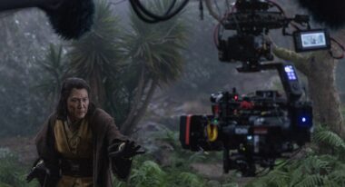 An Asian man in Jedi costume poses in front of a camera on a forest set.