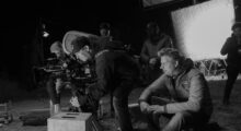 Two white men, one behind a 35mm film camera, on a film set, captured in black and white.