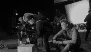 Two white men, one behind a 35mm film camera, on a film set, captured in black and white.