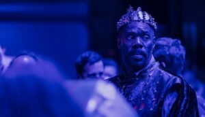 A Black man in a crown stands on stage under a purple light.