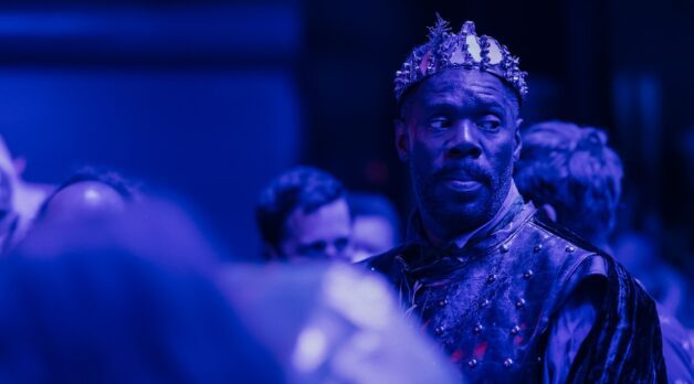 A Black man in a crown stands on stage under a purple light.
