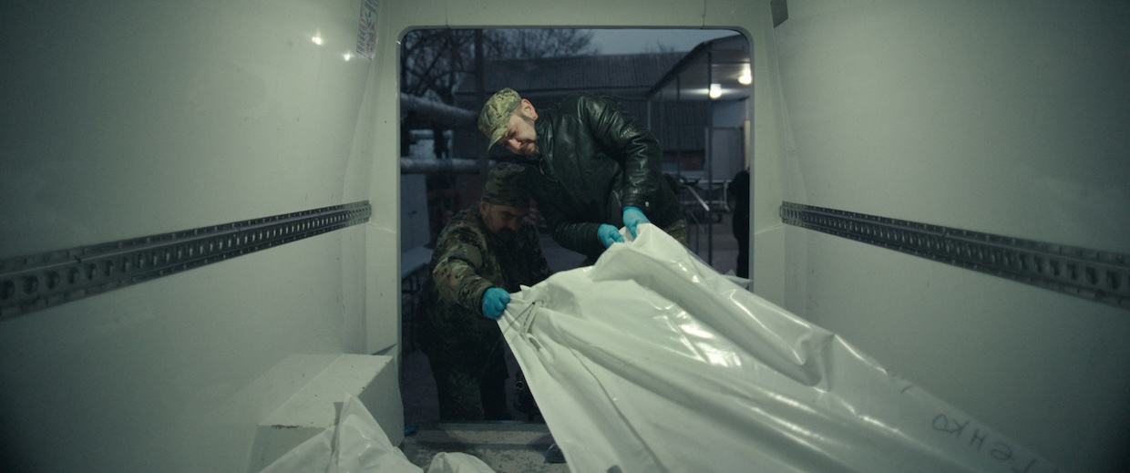 Two men drag a white body bag out of the back of a refrigerated van.