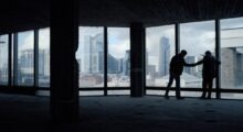 Two people are seen I silhouette shaking hands against the large windows of an upper-floor skyscraper.