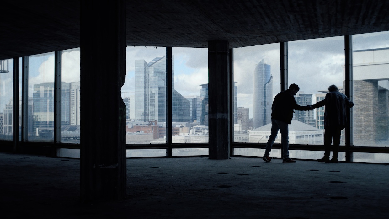 Two people are seen I silhouette shaking hands against the large windows of an upper-floor skyscraper.