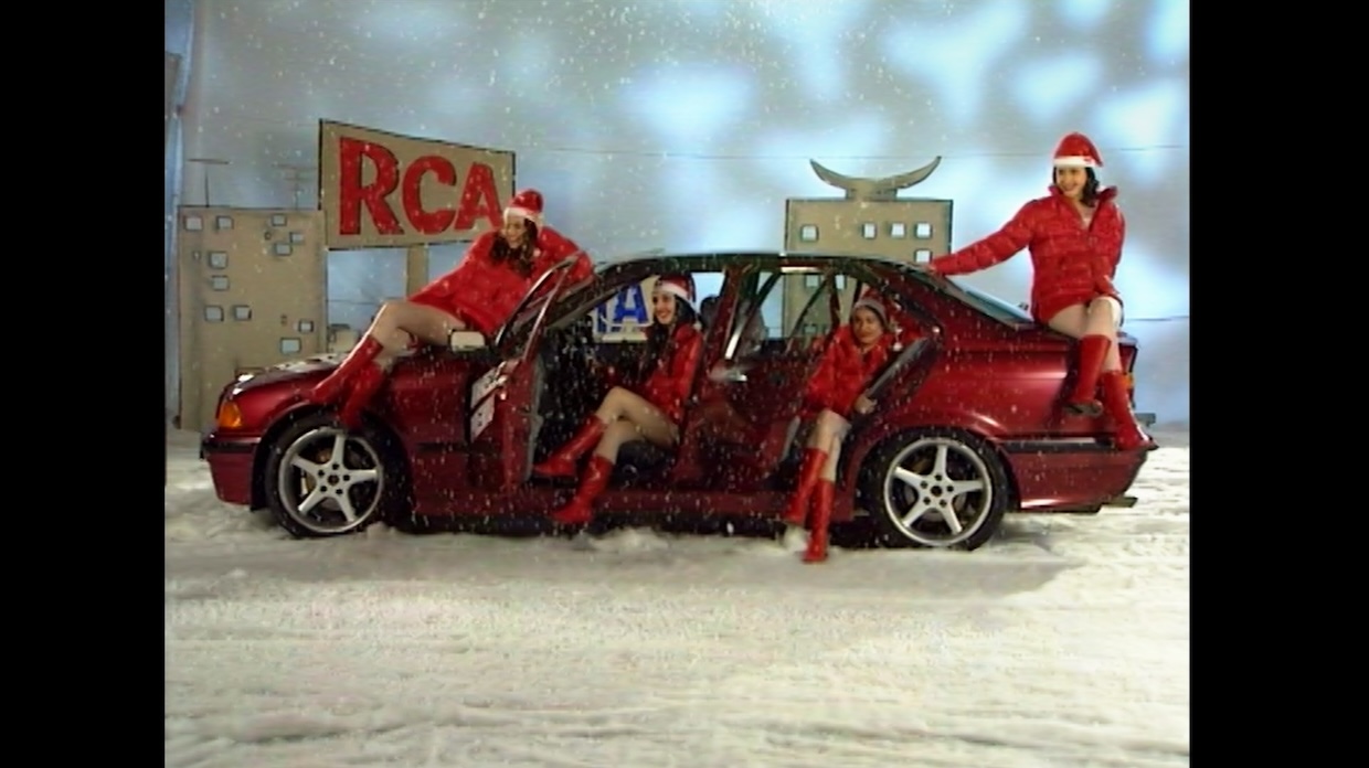 Women in Santa hats and suits sit in a red car on a soundstage with an RCA sign painted behind them.