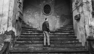 A man stands in the center of two staircases leading left and right outside an old building.