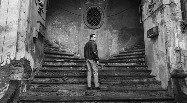 A man stands in the center of two staircases leading left and right outside an old building.