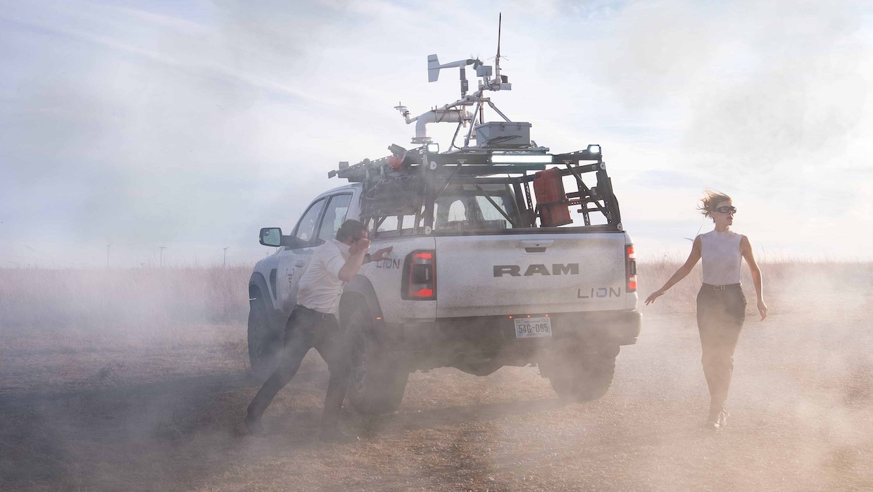 A man and a woman are trapped in a dust storm surrounding their large, custom-modified Dodge Ram.