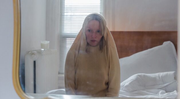 A woman sits on a bed with her sweater pulled over the top of her head.