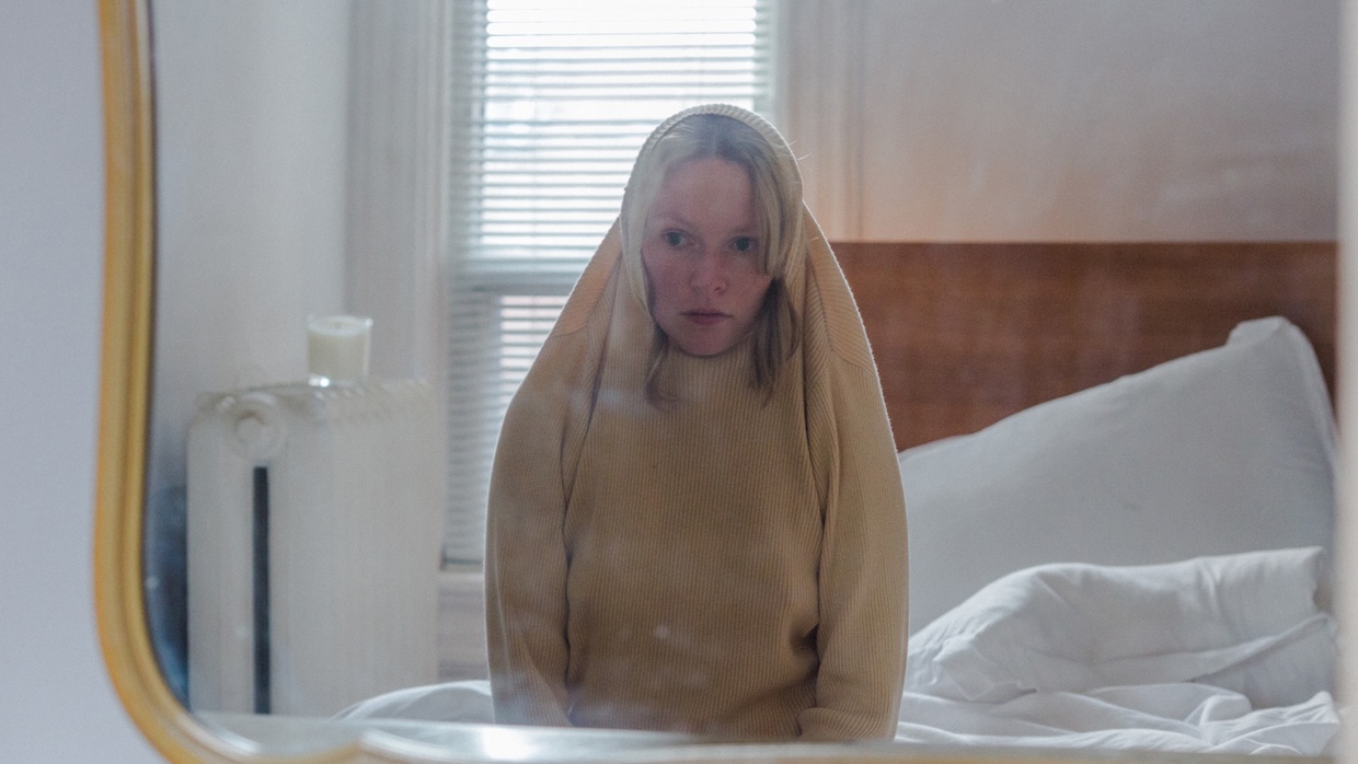 A woman sits on a bed with her sweater pulled over the top of her head.