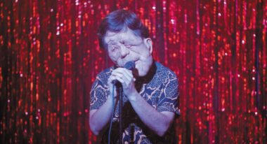 A man with neurofibromatosis sings onstage against a sparkly red curtain.