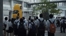 A group of students stare at a yellow car resting vertically on its back end in a courtyard.