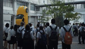 A group of students stare at a yellow car resting vertically on its back end in a courtyard.