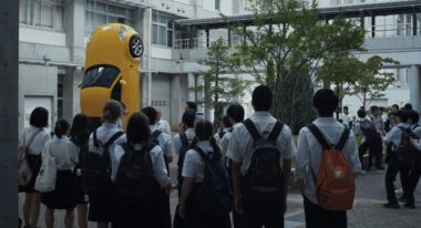 A group of students stare at a yellow car resting vertically on its back end in a courtyard.