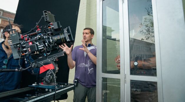 A man with headphones around his neck explains a shot to a camera on a film set.