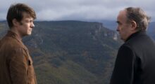 A young man and a priest talk to each other while standing on a rural cliff overlooking a green, hilly landscape.